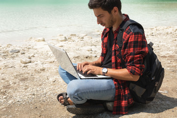 Young brunette male in jeans and shirt is working at a computer while being on the nature. The concept of work on freelancing.