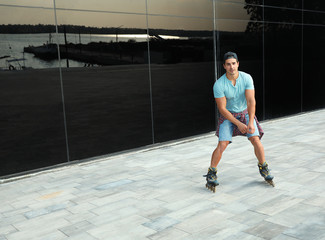 Handsome young man roller skating near glass building on city street, space for text