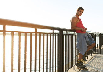 Handsome young man with roller skates on pier near river, space for text