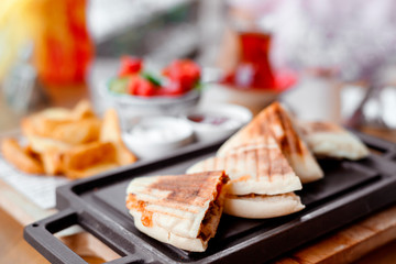 quesadilla sandwich with meat lies on black board on table in outdoor cafe. Turkish tea in glass, potatoes and vegetable salad next to it
