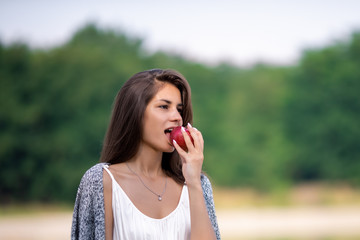 Fototapeta premium Young beautiful girl in white dress eating ripe organic apple. She is on the shore of the lake and enjoy a picnic and outdoor recreation.