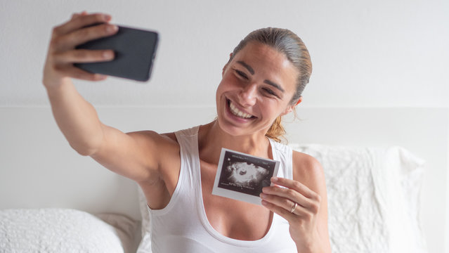 Authentic close up shot of an young pregnant woman in pajamas is sitting on a bed and making a video call to her relatives or friends and showing an ultrasound to announce a future child birth.