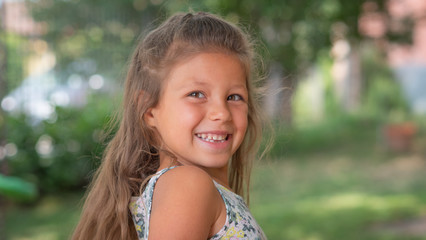 Authentic shot of cute little girl with loose hair is smiling in camera outside her house on a sunny day.