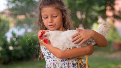 Authentic moment of happy little smiling girl is holding a white hen outside the countryside house in a sunny summer day.