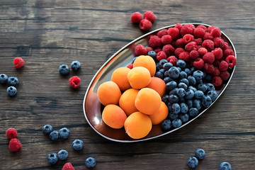Fresh apricots, raspberries and blueberries on an oval metal plate. Gray background.