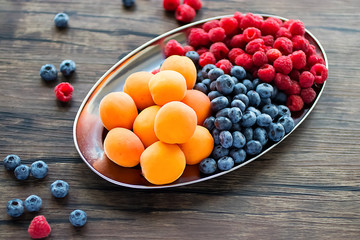 Fresh apricots, raspberries and blueberries on an oval metal plate. Gray background.