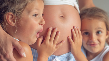 Authentic shot of two lovely little daughters are touching mother's pregnant belly, feeling baby at mother tummy and awaiting the birth of their little brother or sister.
