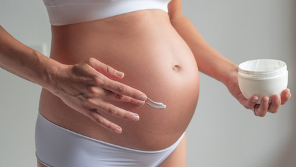 Authentic close up shot of an young pregnant woman in cosy underwear is applying a moisturizer or...