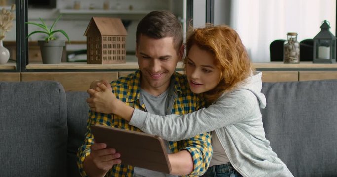 Young man and woman found a product they like in online store, using their tablet, woman is hugging man, being surprised, saying wow, smiling, sitting on a couch. 4K, shot on RED camera.