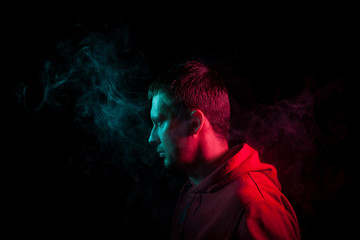 Portrait of a guy in profile face standing back to black background with a feeling of sadness and loneliness, around his head a cloud of blue and pink smoke. The soul and feelings of man