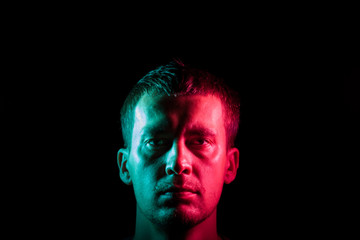 Close-up of the face of an unshaven face of a caucasian man with stubble in the dark, illuminated on one side in Blue and on the other pink on a black isolated background