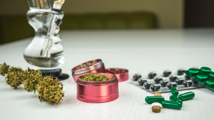 Marijuana buds, bong, grinder and pills lie on the white table.