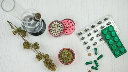Marijuana buds, bong, grinder and pills lie on the white table.