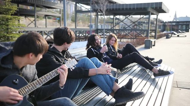 Young friends chilling on a bench - drinking alcohol and playing the guitar