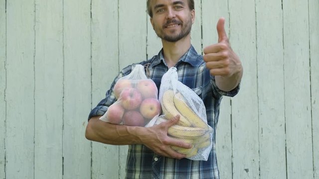 Zero waste shopping concept. Hipster man holding reusable eco bags with fresh fruits and showing LIKE sign. Ban single use plastic.