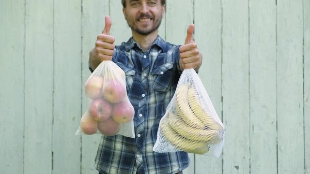 Happy smiling adult man holding reusable eco bags with fresh fruits and showing LIKE sign. Zero waste shopping, ban single use plastic concept.