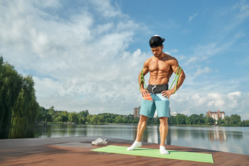 Muscular man with rubber expander posing near lake.