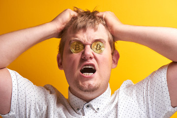 Portrait of upset fat man with bitcoins in eyes. Guy tears hair on head, yells, put money into cryptocurrency and lost everything on yellow background