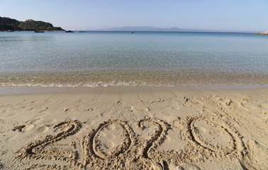 Happy New Year 2020, lettering on the beach with wave and clear blue sea. Numbers 2020 year on the sea shore, message handwritten in the golden sand on beautiful beach background. New Years concept.