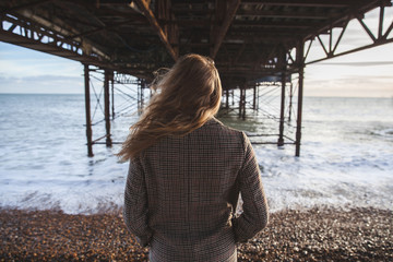 Blond young Woman standing under the bridge in coat looking towards the ocean. Moody landscape. Brighton, West Sussex, England 