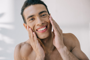 handsome mixed race man touching cheeks with hands, smiling and looking at camera