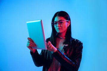 Beautiful female half-length portrait isolated on blue studio background in neon light. Young emotional woman. Human emotions, facial expression concept. Using tablet for talking or internet serfing.