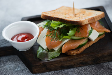 Sandwich with salmon fillet, cheese, salad leaves and cucumber on a black wooden serving tray, selective focus
