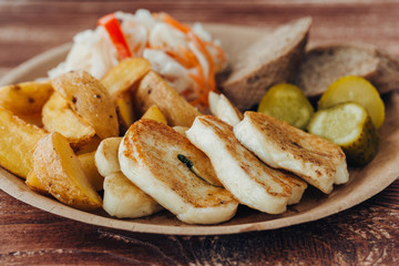 Tasty grilled fish with fried potatoes, cucumber, cabbage and woods in a paper plate on a wooden table. takeaway street food