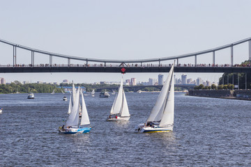 Fototapeta premium Yachts with white sails sailing on the river. Over the river bridge with pedestrians