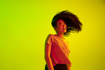 Beautiful female half-length portrait isolated on yellow studio background in neon light. Young emotional woman. Human emotions, facial expression concept. Dance in eyeglasses and wireless headphones.