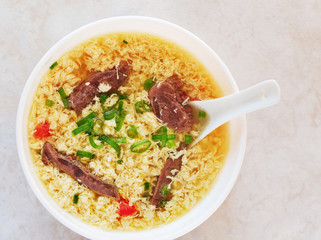 Egg drop soup also known as egg flower soup made of beaten eggs drizzled in homemade bone broth with pieces of meat. Top view of soup in white bowl.