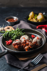 Grilled chicken shins in tomato sauce with chili pepper on a dark pan and baked potatoes in a bowl