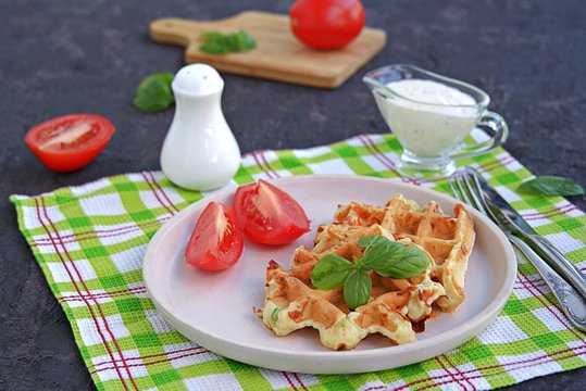 Savory zucchini waffles on a white plate on a dark concrete background. Served with ranch dressing and fresh tomato. Healthy food.