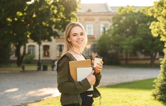 Young smiling female student holding take away coffee in the park near the college. Pretty, cute casual student near the high school building with books and coffee. Park, green lawn.