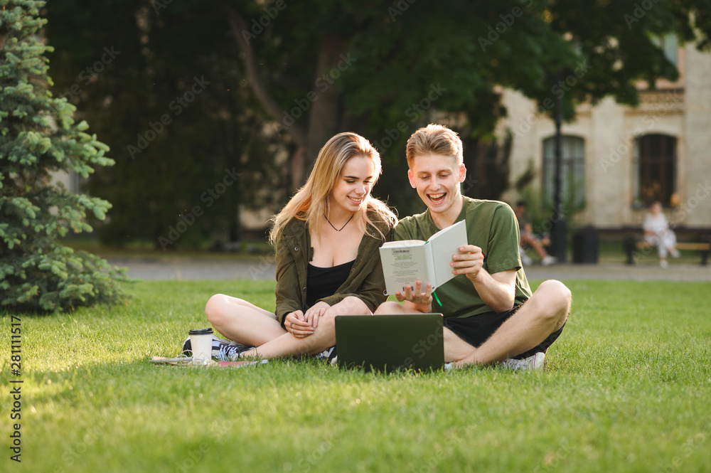 Wall mural Couple of attractive smiling students dressed casually studying outdoors on campus lawn at the university. Two young smiling students reading the book sitting with laptop on grass in the college park.