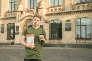 Portrait of serious caucasian male student wearing green T-shirt standing in front of university holding copybooks and coffee. Concentrated calm student standing in college yard with coffee and books.