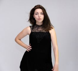 Fototapeta premium A portrait above the knee of a pretty beautiful fashionable adult brunette girl in a black dress on a white background. Standing right in front of the camera, showing different poses and emotions.