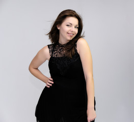 Fototapeta premium A portrait above the knee of a pretty beautiful fashionable adult brunette girl in a black dress on a white background. Standing right in front of the camera, showing different poses and emotions.