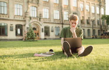 A thoughtful guy sitting on the lawn on the background of the university with a laptop, thoughtfully looking at the screen and studying. Portrait of a young student working with a laptop in campus.