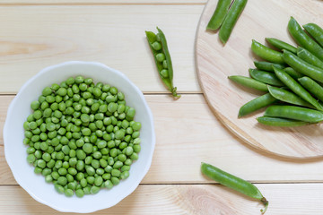 Green peeled peas in white plate on light wooden table with whole pods on round kitchen desk