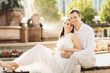 Young couple sitting on a bench at the fountain. A man gently embraces his pregnant wife. Germany,...