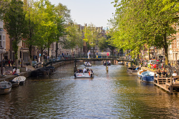 Obraz premium Amsterdam, Netherlands - 06/14/2019: canal with bridge and boats in Amsterdam, Netherlands. Traditional dutch cityscape. Historic street in Amsterdam with boats and tourists. Summer travel concept.