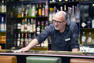 Elderly man bartender wiping the table with a rag at the hotel bar. Bartender in the workplace. Shelves with bottles of alcohol in the background. The concept of work and service.