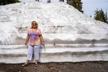 Cute blonde tourist woman poses and stands next to a large mound of plowed snow in Lassen National...