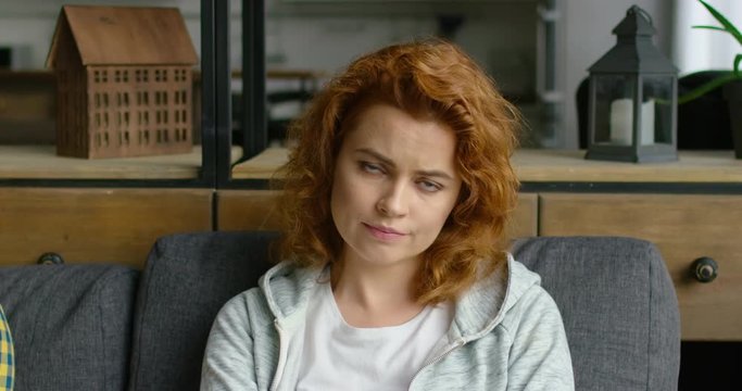 Portrait of a young annoyed woman sitting on a sofa at home, angry, looking at the camera, with red curly hair, blue eyes, Caucasian. 4K, shot on RED camera.