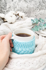 Fototapeta na wymiar A female hand is holding a white mug of tea in a knitted blue frame and wrapped in a white knitted plaid. In the background, sprigs of eucalyptus and cotton. Breakfast in bed. Cozy.