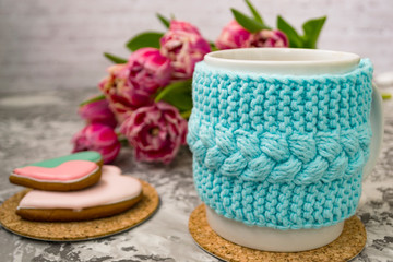 White cup of tea in a knitted blue case with pink flowers tulips and sweets, with gingerbread on a textural table.