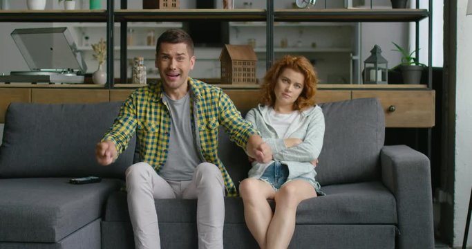 Man and woman are sitting on a couch at home, watchning TV, man is cheering for his favorite sport team, yelling and excited, hugging woman that is borred and uninstersted. 4K, shot on RED camera.