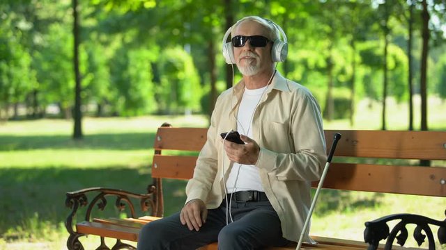 Blind old man wearing earphones listening audiobook, voice message in cellphone