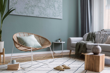 Modern and bohemian composition of interior design with gray sofa, rattan armchair, wooden cube,...
